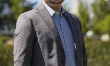 A bald man with a beard and glasses is smiling while standing outdoors. He is wearing a gray suit jacket over a light blue dress shirt. The background features green trees and a blurred modern building under a clear blue sky.
