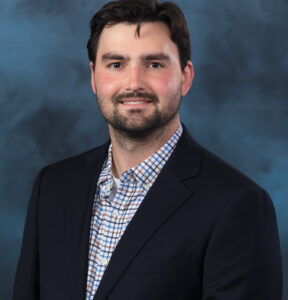 A man with dark hair and a short beard wearing a navy blazer over a plaid button-down shirt smiles in front of a blue mottled studio backdrop.
