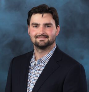 A man with dark hair and a short beard wearing a navy blazer over a plaid button-down shirt smiles in front of a blue mottled studio backdrop.