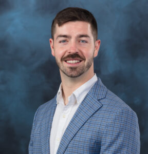 A man with short brown hair, a mustache, and a beard wearing a light blue checkered blazer over a white button-down shirt smiles in front of a blue mottled studio backdrop.
