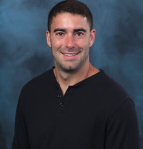 A man with short dark hair wearing a black henley shirt smiles in front of a dark blue mottled studio backdrop.