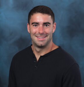 A man with short dark hair wearing a black henley shirt smiles in front of a dark blue mottled studio backdrop.