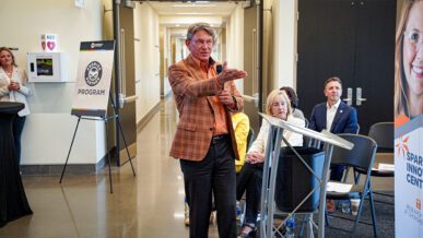 A man in an orange plaid jacket speaks while gesturing with his hand at a Premier Partner’s event, hosted at the Spark Innovation Center. Several people are seated and listening in the background, with event signage visible.