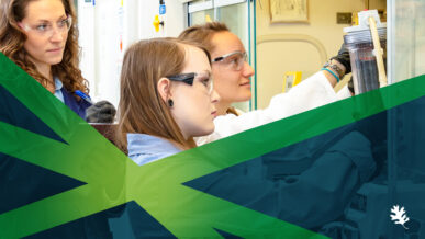 Three people wearing safety glasses and lab coats work together at a laboratory station. One person is adjusting equipment inside a fume hood while the others observe. A green and blue graphic overlay with an oak leaf logo runs across the bottom of the image.