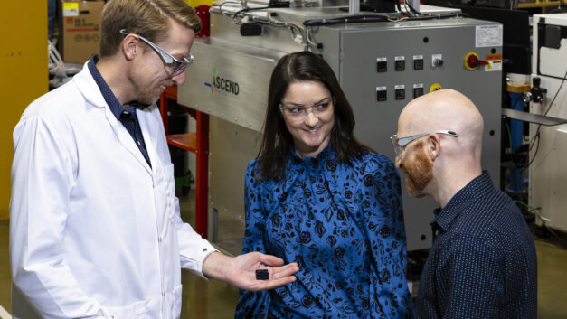 Three people wearing safety glasses stand together in a manufacturing facility. A man in a white lab coat on the left is holding a small 3D-printed part in his hand and showing it to the other two. A woman in a blue patterned blouse smiles while looking at the part, and a bald man with a red beard observes from the right. Large industrial equipment with the Ascend Manufacturing logo is visible in the background.