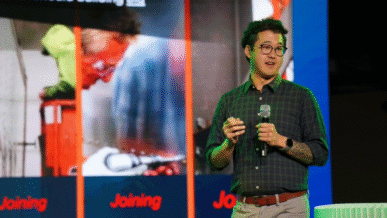 Daniel Lee, founder and CEO of Perseus Materials, presenting on stage during a pitch event. He gestures toward a screen behind him while speaking to an audience, highlighting his company’s innovative composite manufacturing technology.
