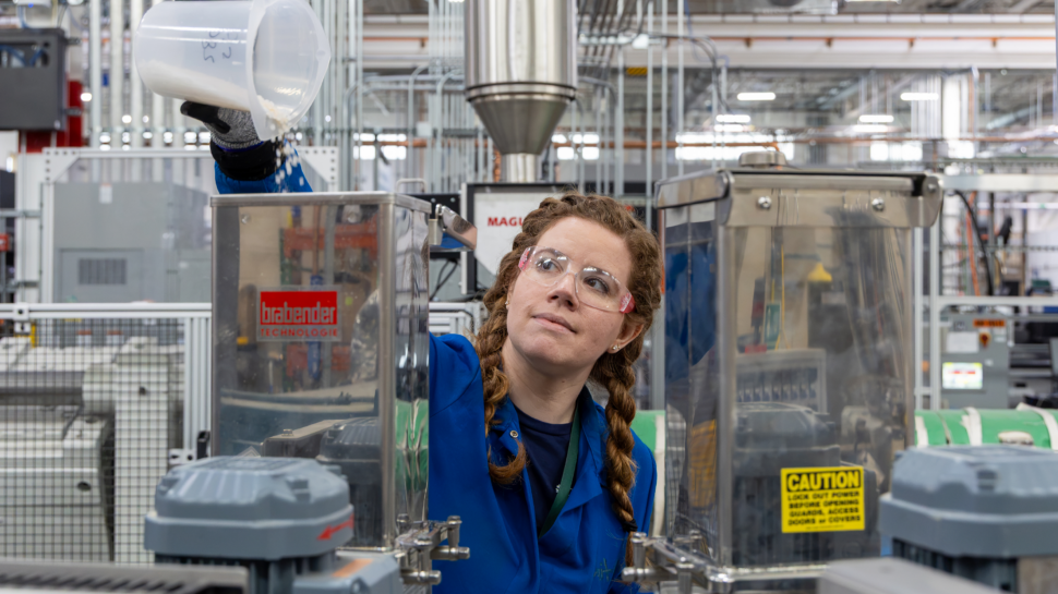 Cait Clarkson, wearing safety glasses, gloves and a blue lab coat, pours white polymer pellets from a container into the hopper of plastics-processing equipment.